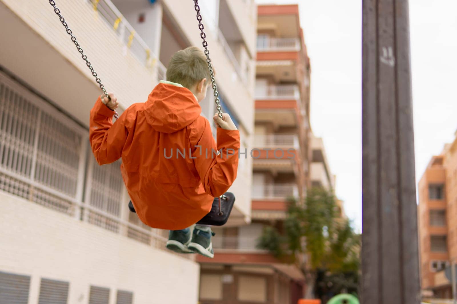 a boy with blond hair in an orange jacket rides on a swing turned with his back to the camera on the street. There is a place for an inscription by PopOff