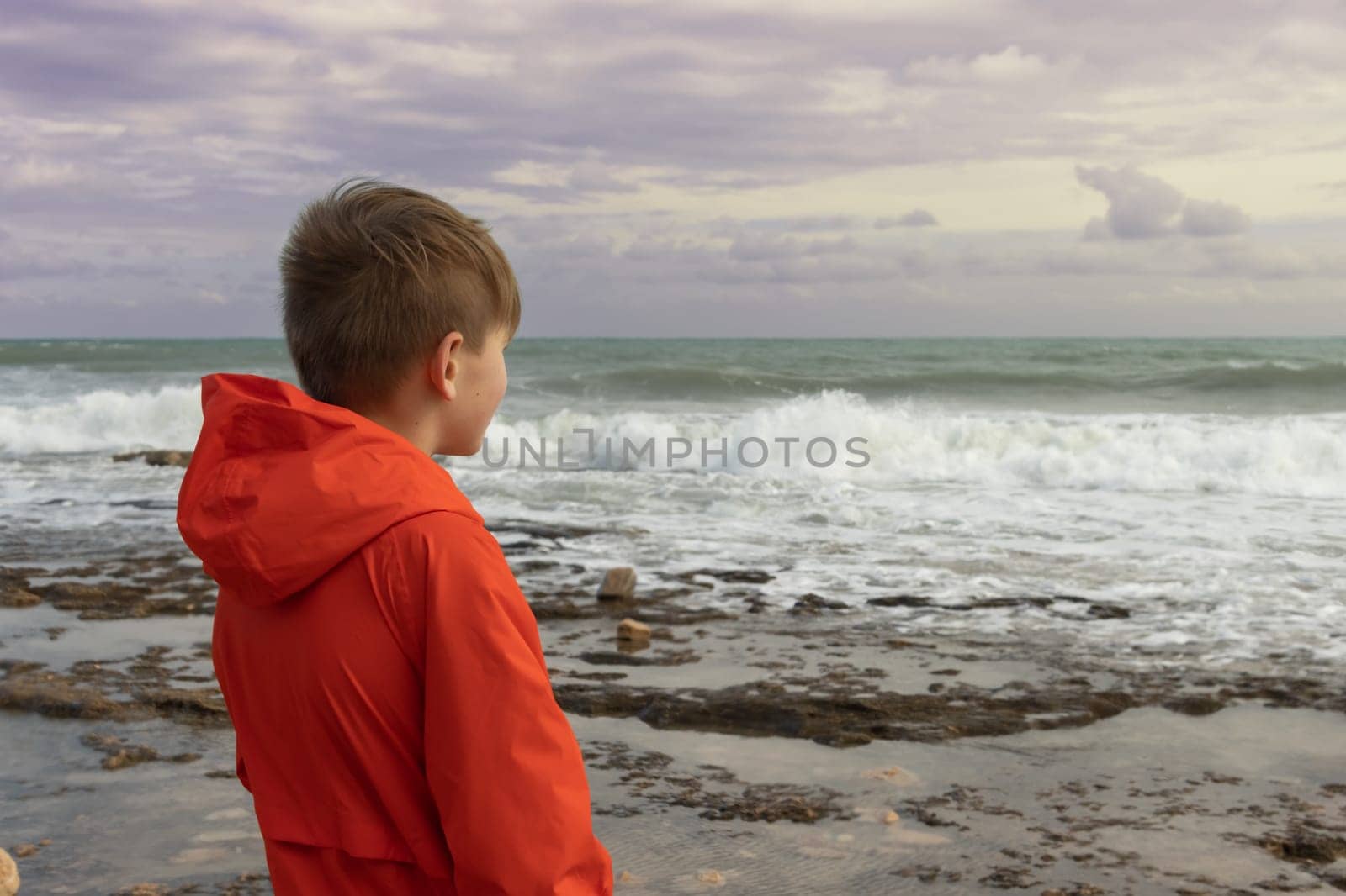 a boy of European appearance with blond hair in an orange windbreaker, standing on the embankment close-up, there is a place for an inscription by PopOff