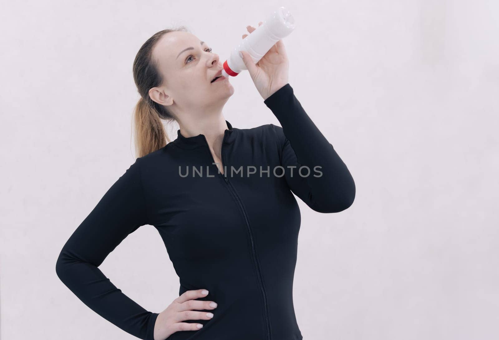 Portrait of a young tired caucasian fitness woman standing in black sportswear and drinking water from a white bottle isolated on white background. Home workout concept