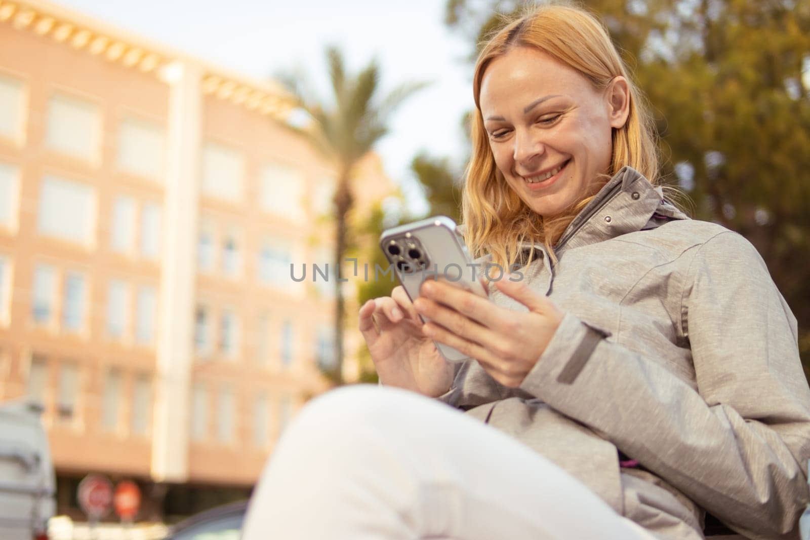 a smiling girl of European appearance with blond hair sits on a bench, looks at the phone and smile by PopOff