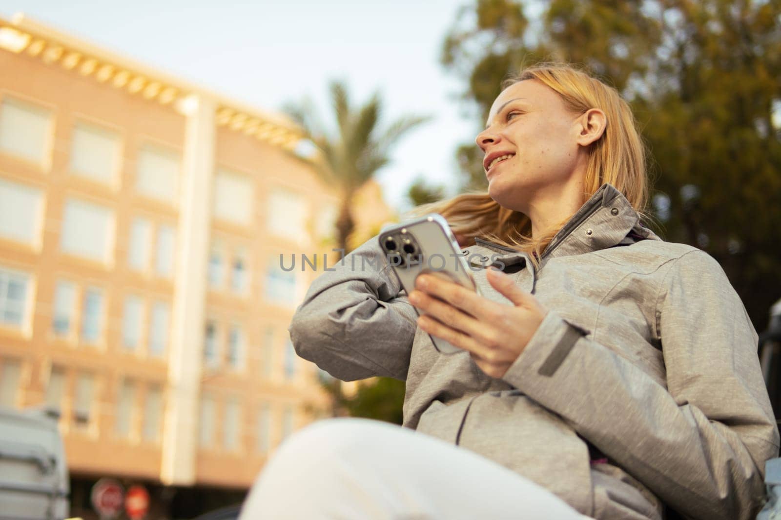 a pensive girl of European appearance with blond hair sits on a bench, holds a phone in her hands and looks sideways smiling, a girl sits in a gray jacket and white sweatpants. High quality photo