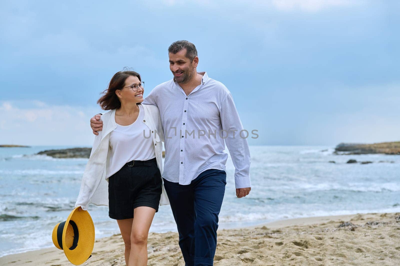 Happy mature couple walking on the beach. Smiling talking middle-aged man and woman embracing together on the seashore. Age, relationship, love, leisure, vacation, people concept