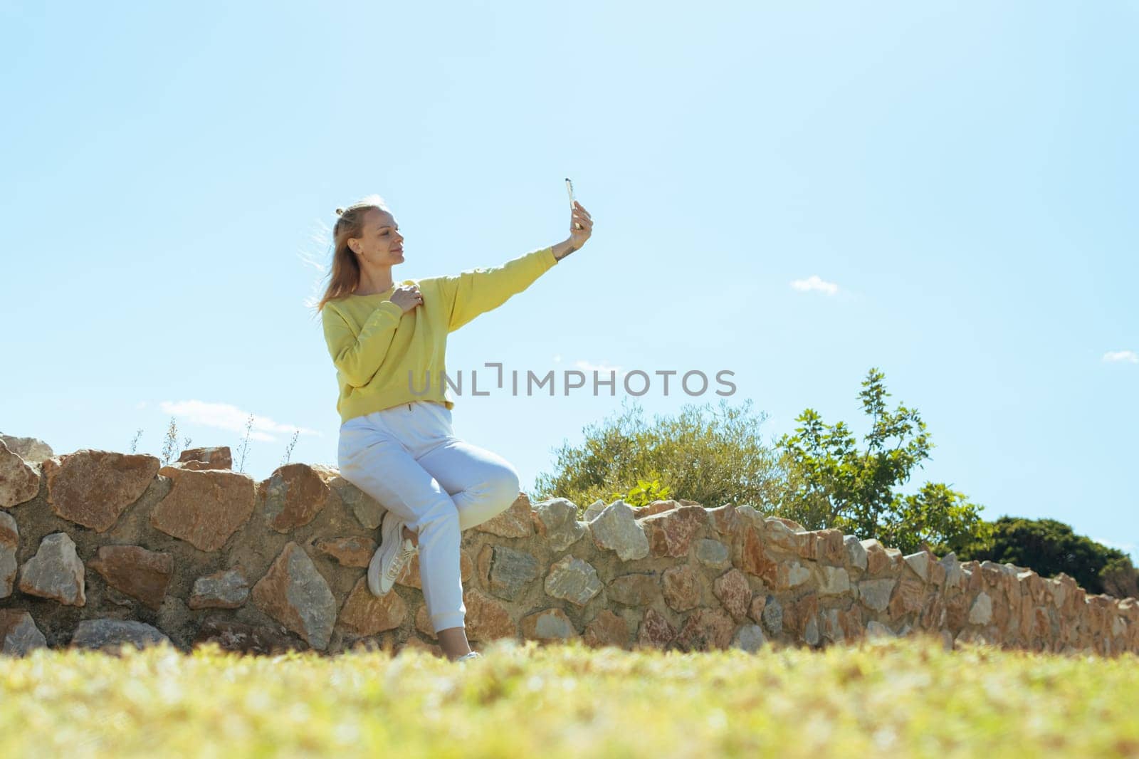 a beautiful girl sits on a fence with stones and takes a selfie against the blue sky .beautiful landscape with a girl by PopOff