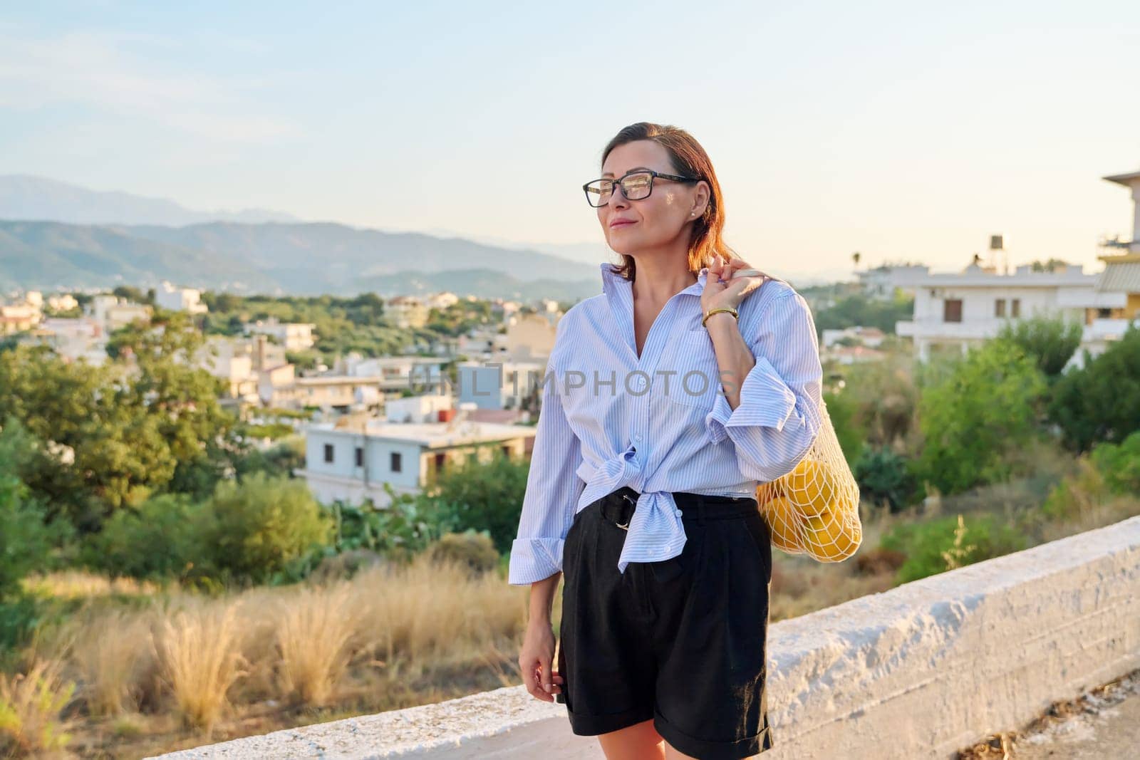 Eco tourism, woman walking relaxing in European mountain village, enjoying clean air, landscape, sunset, with a bag of fresh farm oranges. Nature, travel, ecology, lifestyle, people concept