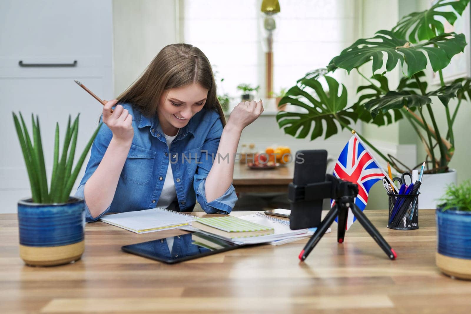 Online English lesson, young woman teaching English to students, using smartphone on tripod. Virtual lesson, teacher teaching remotely, recording video lesson.