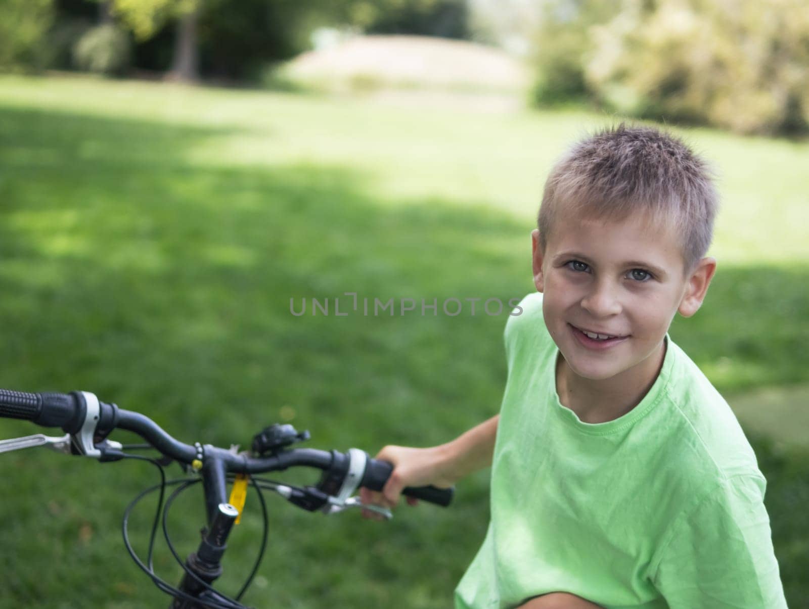 a teenager boy of European appearance with short blond hair in a green T-shirt stands with a bicycle in the park.looks at the camera on the left there is a place for an inscription. High quality photo
