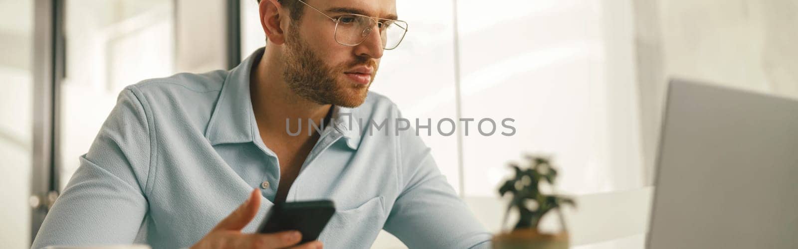 Male entrepreneur holding phone while working on laptop in modern office. High quality photo