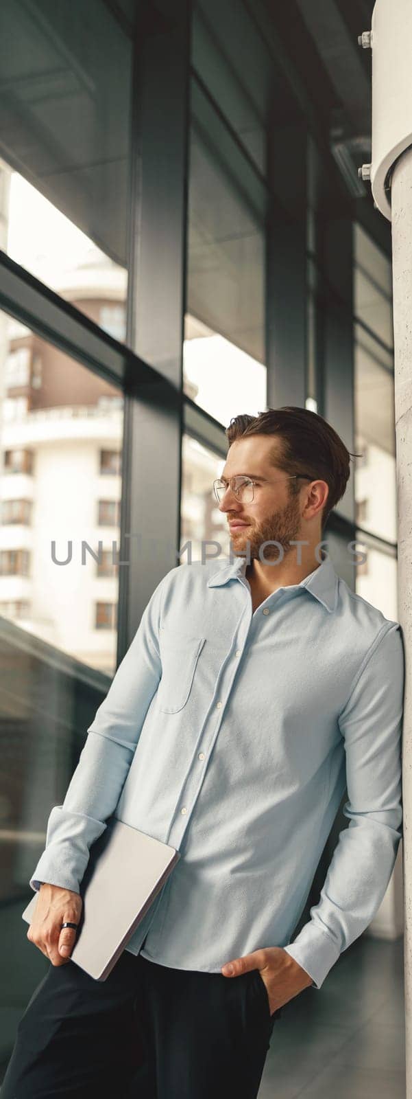 Stylish businessman in eyeglasses with laptop standing on office background and looking at side