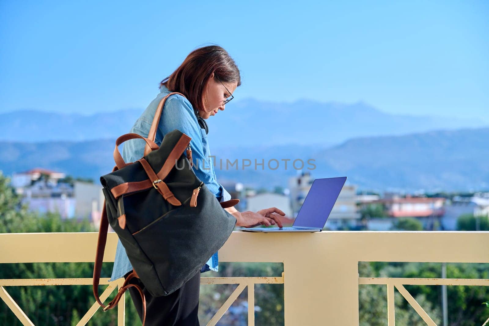 Confident serious mature businesswoman with laptop outdoors. Middle-aged female in denim shirt headphones glasses with backpack looking at laptop screen. Work, business, technology, people 40s age