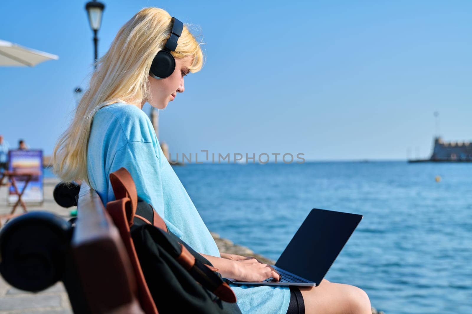 Young woman relaxing in headphones with a laptop on the seashore, in a touristic summer city. Beautiful teenage blonde enjoying her vacation. Youth, vacation, technology, tourism concept