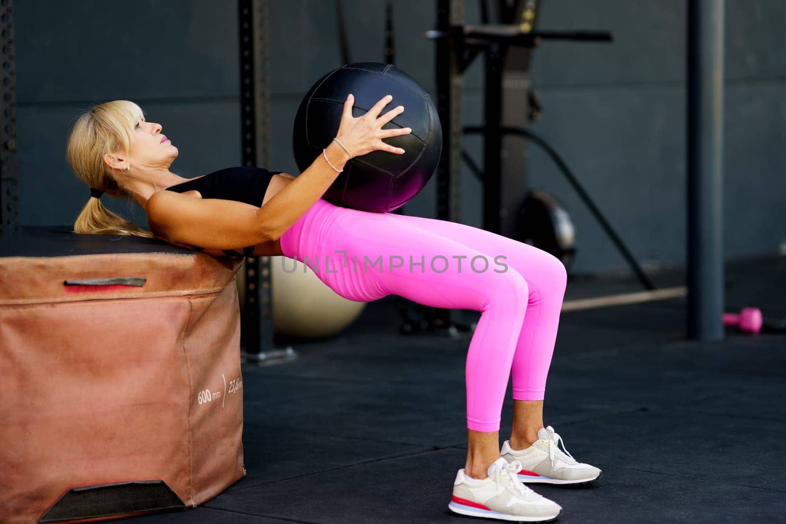 Side view of strong fit female doing exercise with heavy ball while exercising against fitness equipment in modern gym during workout and looking up