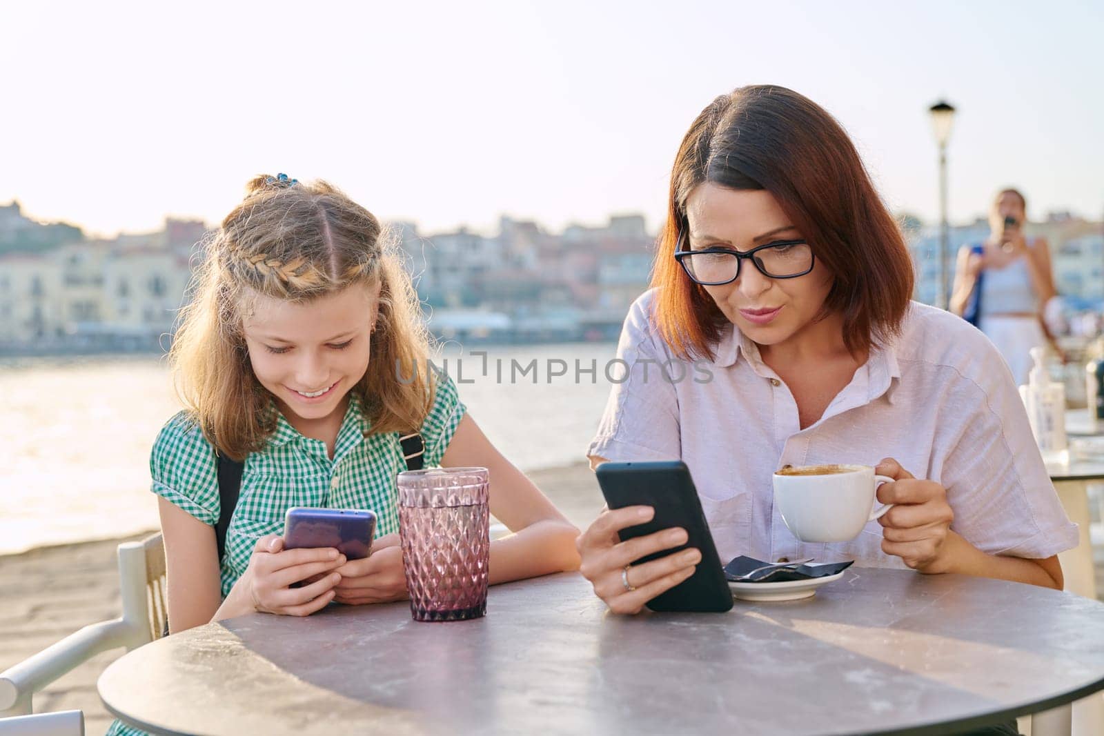 Mother and daughter child together in an outdoor cafe on the waterfront. Family, mom and preteen girl using smartphones, with coffee, drinks. Lifestyle, family, parent and child concept