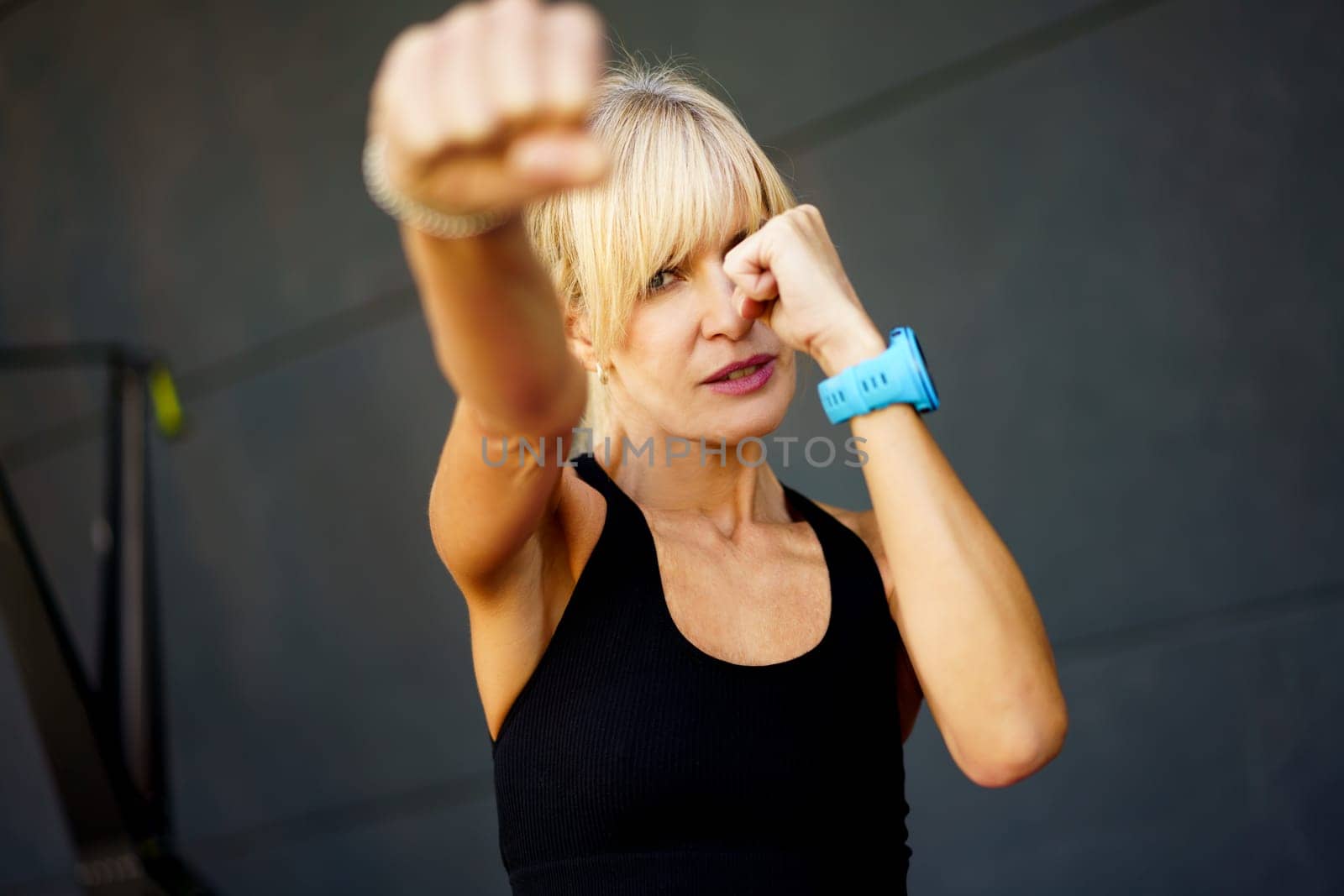 Confident young fit blond haired female wearing black tank top performing martial art punch in fitness gym against gray background looking at camera