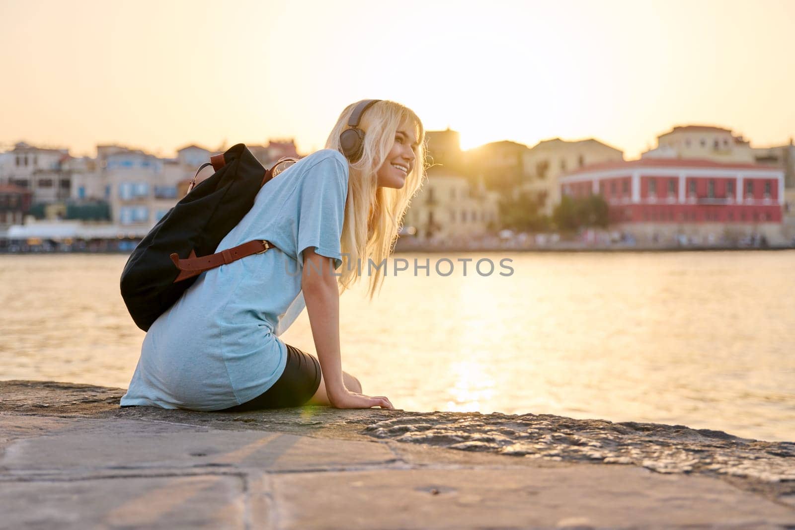 Happy having fun young teenage female in headphones with backpack enjoying old tourist European city, seascape, copy space. Tourism, Greece Crete Chania, summer, vacation, youth, rest, nature, sunset