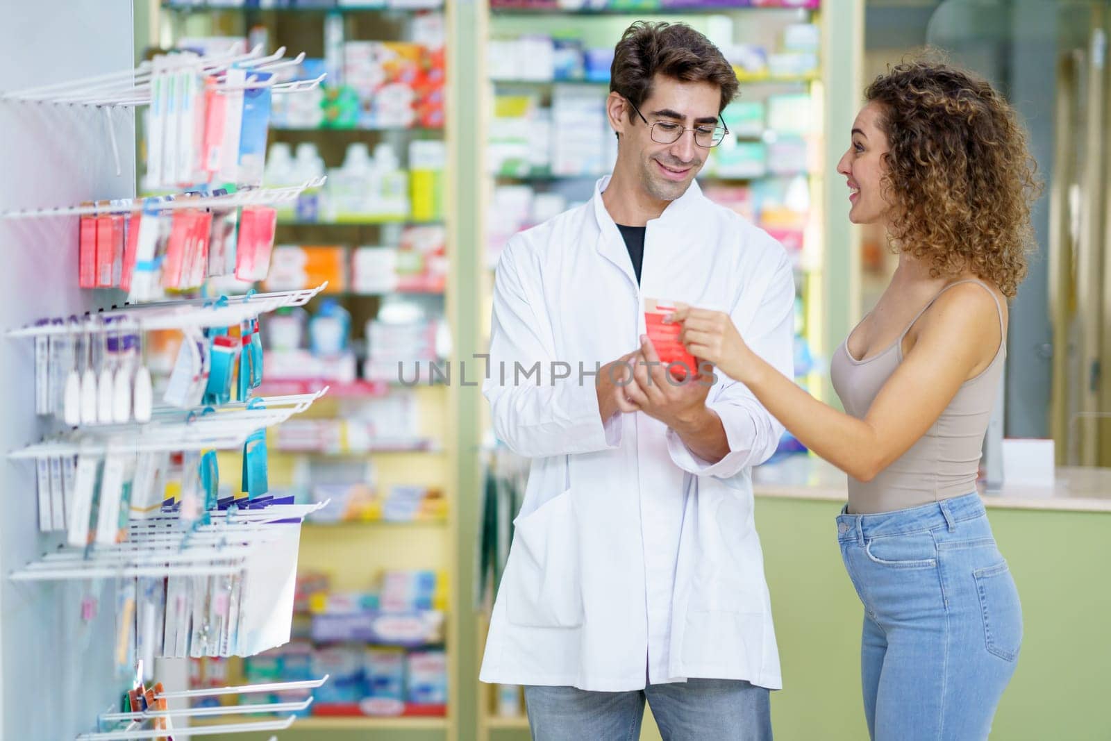 Happy young male pharmacist, in white robe and eyeglasses standing with smiling female customer near shelves with assorted medical supplies and explaining medication details in pharmacy