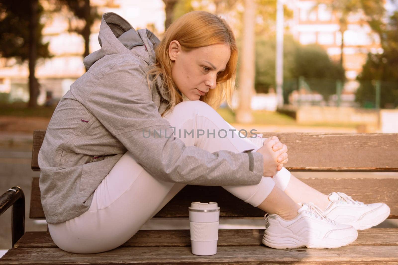 a pensive girl of European appearance with blond hair sits on a bench, a girl sits in a gray jacket and white sweatpants and sneakers. High quality photo