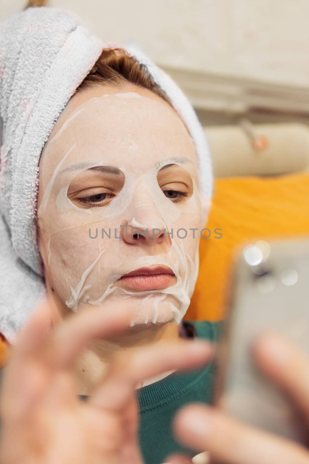 a girl of European appearance lies on a bed with a towel on her head and a mask on her face, doing procedures for facial rejuvenation, flipping through the phone. Relax for a girl. High quality photo