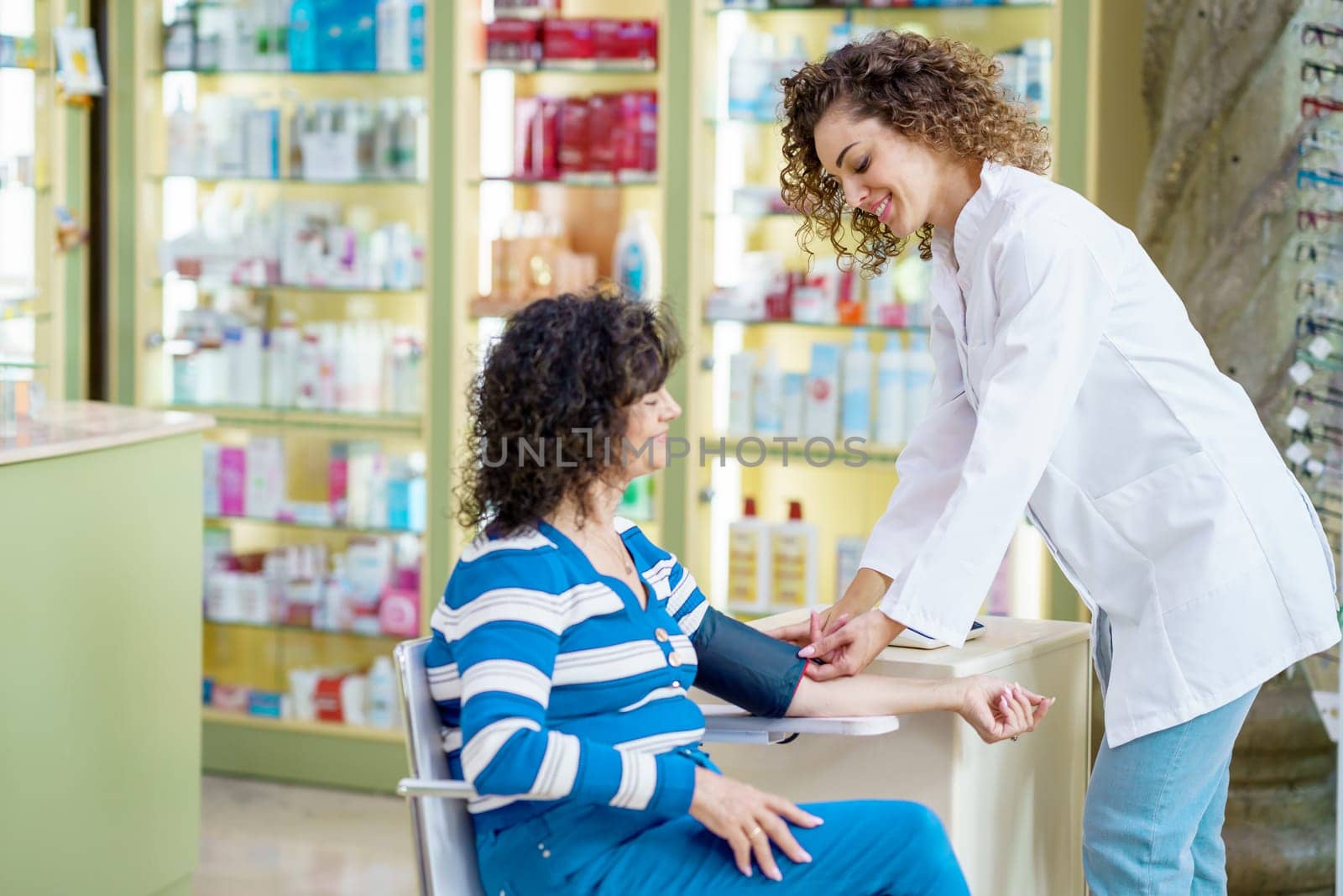 Smiling young female pharmacist in white robe, standing near sitting lady customer and looking at each other during blood pressure checking near shelves with medical supplies in pharmacy