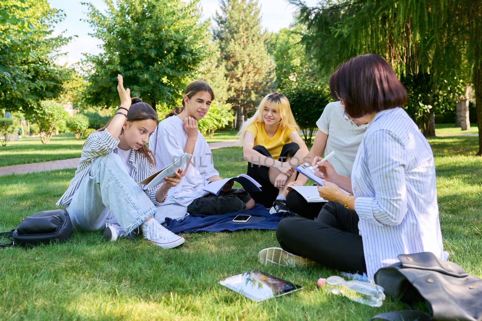Outdoor, group of students with female teacher. Teenagers and mentor teacher talking sitting on grass in college park. Back to school, back to college, high school, education, teenagers concept