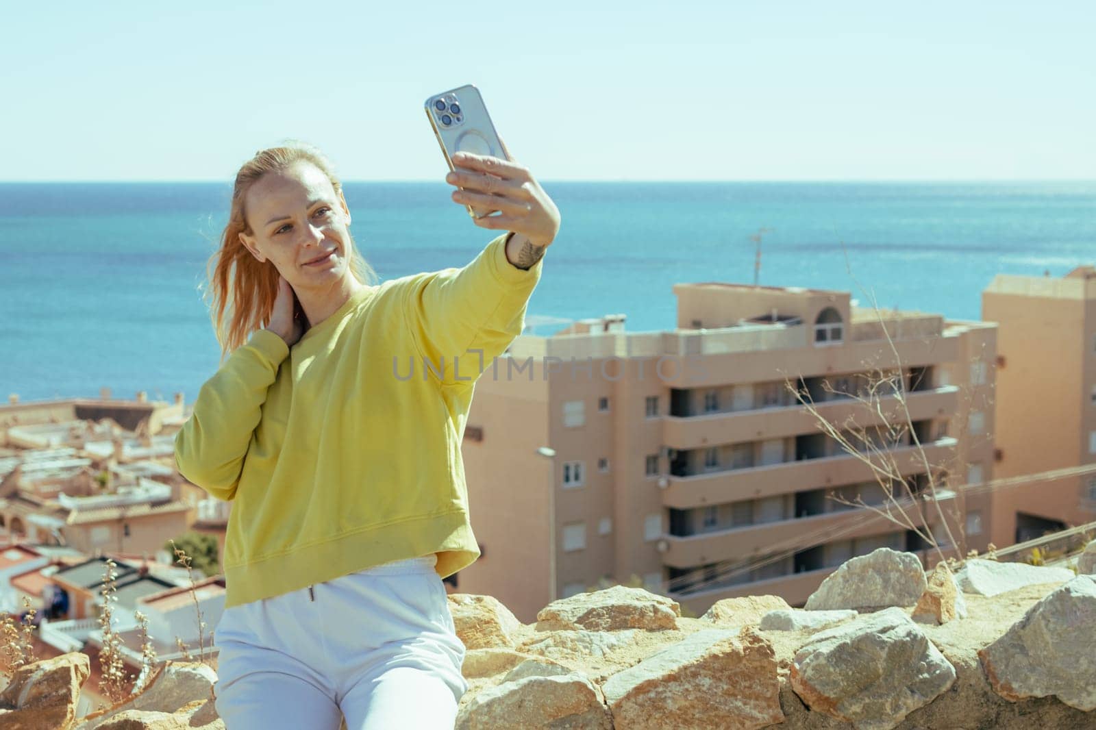 A girl of European appearance is standing in white pants and a yellow jacket taking a selfie overlooking the sea by PopOff