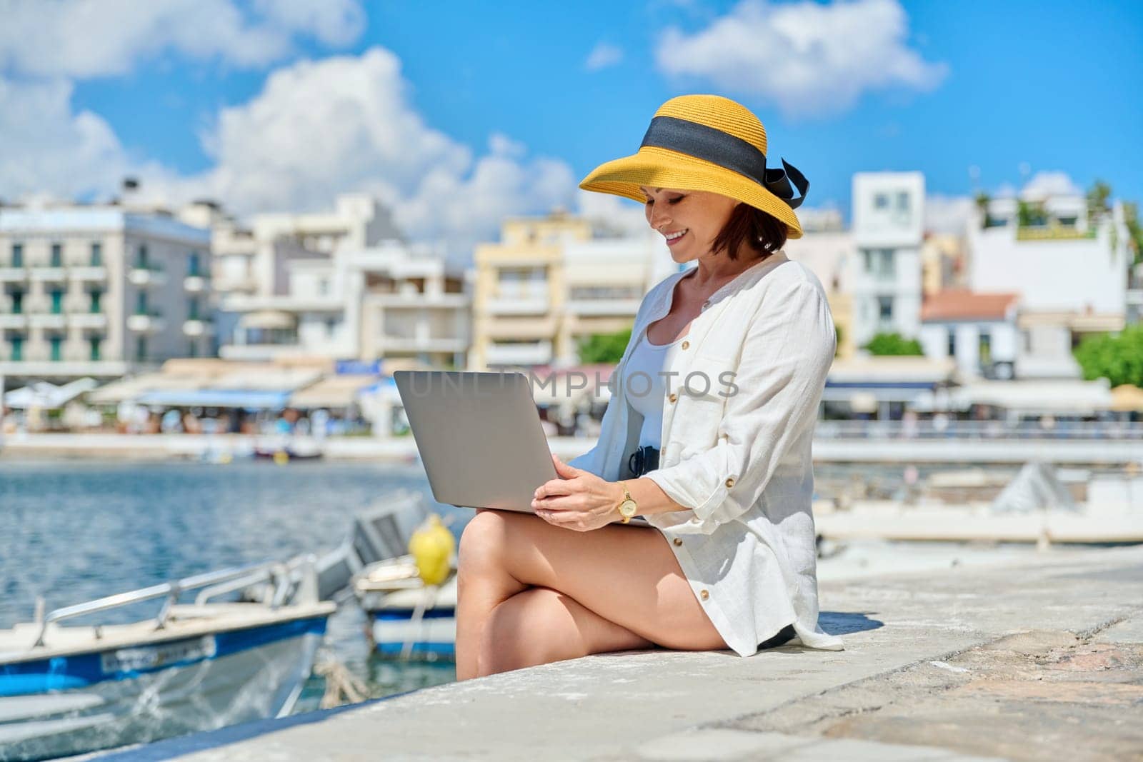Mature woman in hat with laptop on sea promenade in city by VH-studio