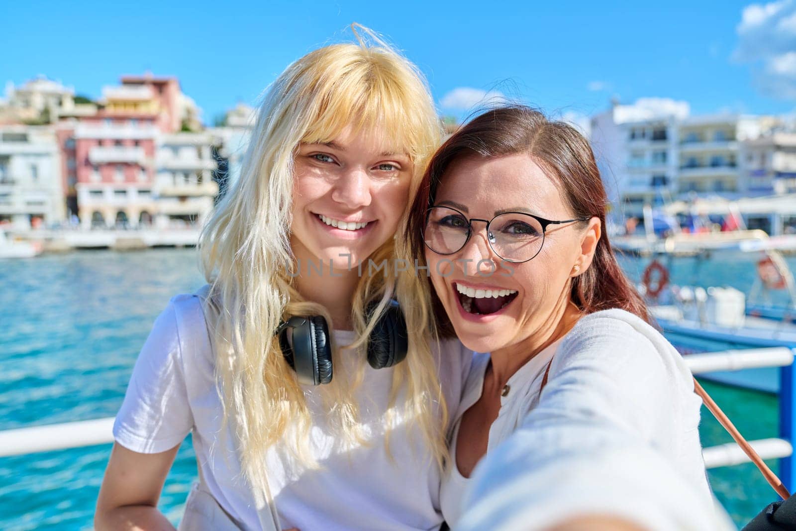Portrait of happy smiling mom and teenage daughter looking at camera together, faces close-up. Sea bay in old tourist town, sunny summer day. Relationship parent teenager, lifestyle, family vacation