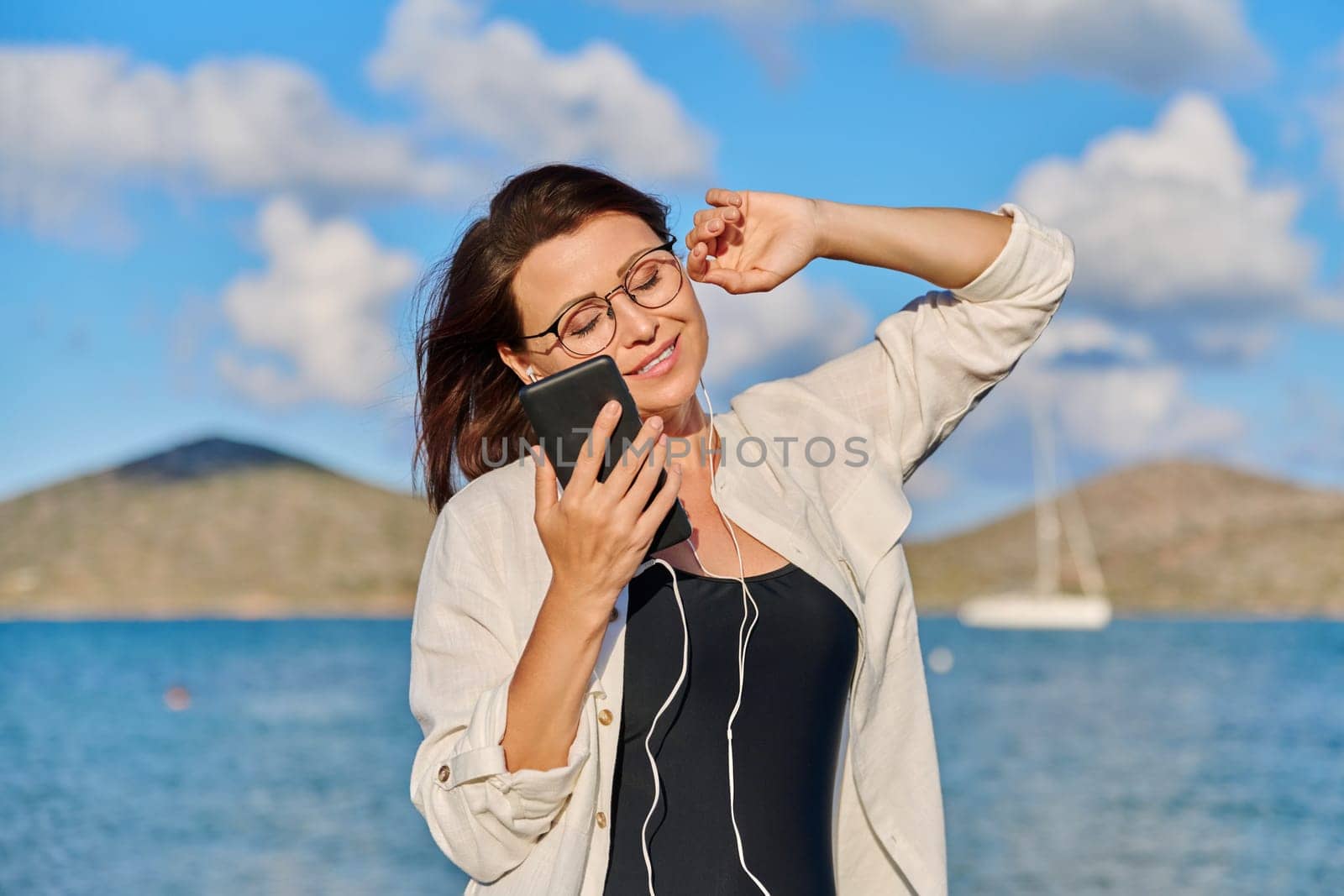 Happy middle aged woman in headphones listening to music using smartphone on beach. Mature female with closed eyes enjoying music, sunset, sea holidays. Vacation, feelings, nature, people 40s concept