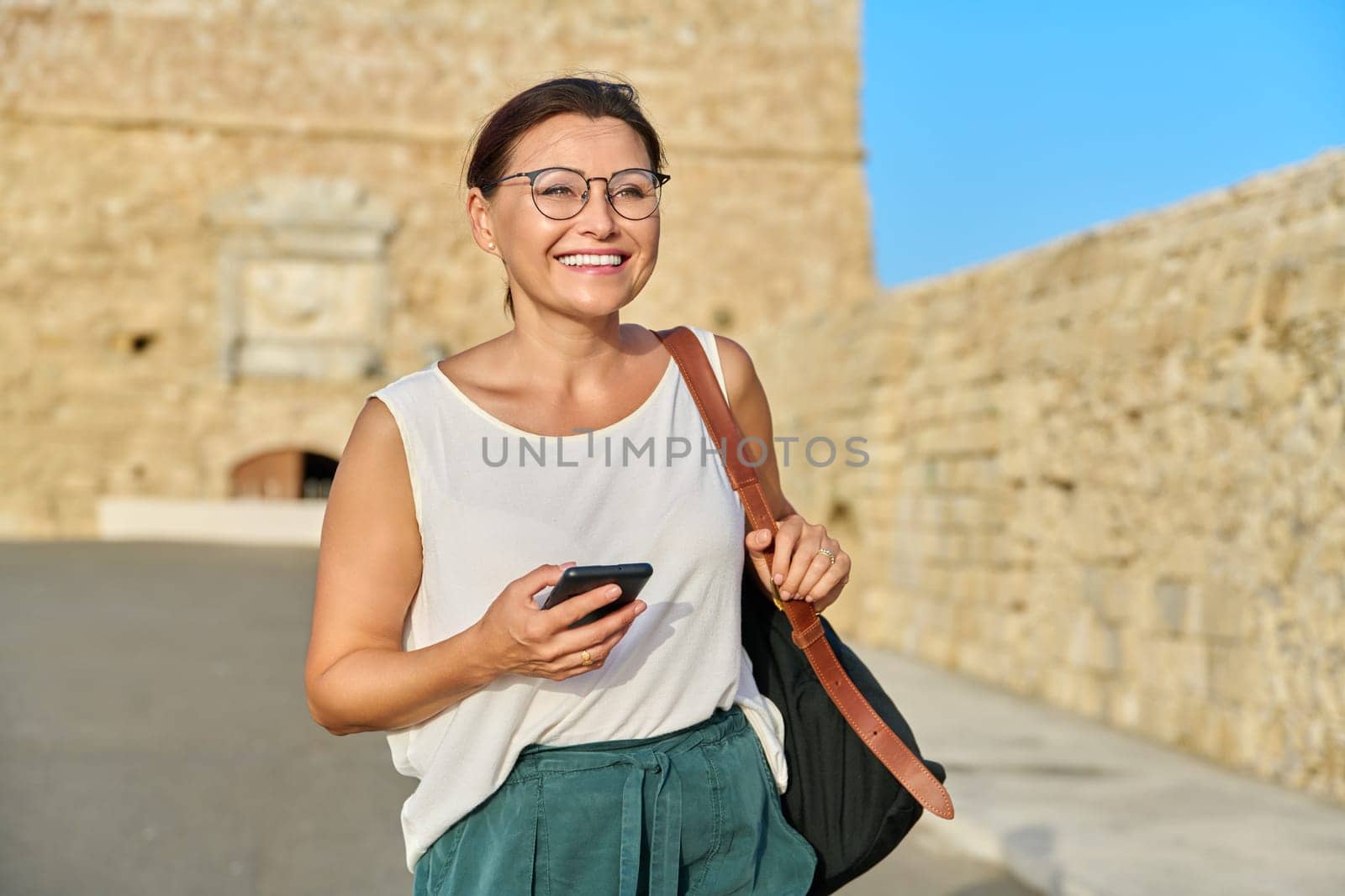 Outdoor portrait of smiling middle-aged woman walking with smartphone in backpack through an old tourist town. 40s age, beauty, health, active lifestyle, travel tourism concept