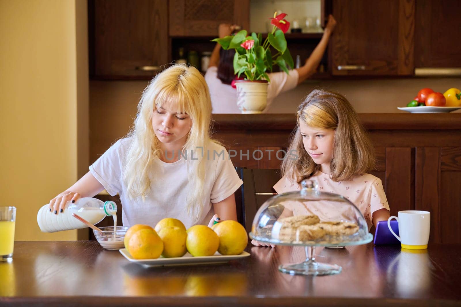 Children two sisters eating at home in the kitchen, teenage and preteen girls having breakfast together, mother cooking. Family, people, children, food, lifestyle concept