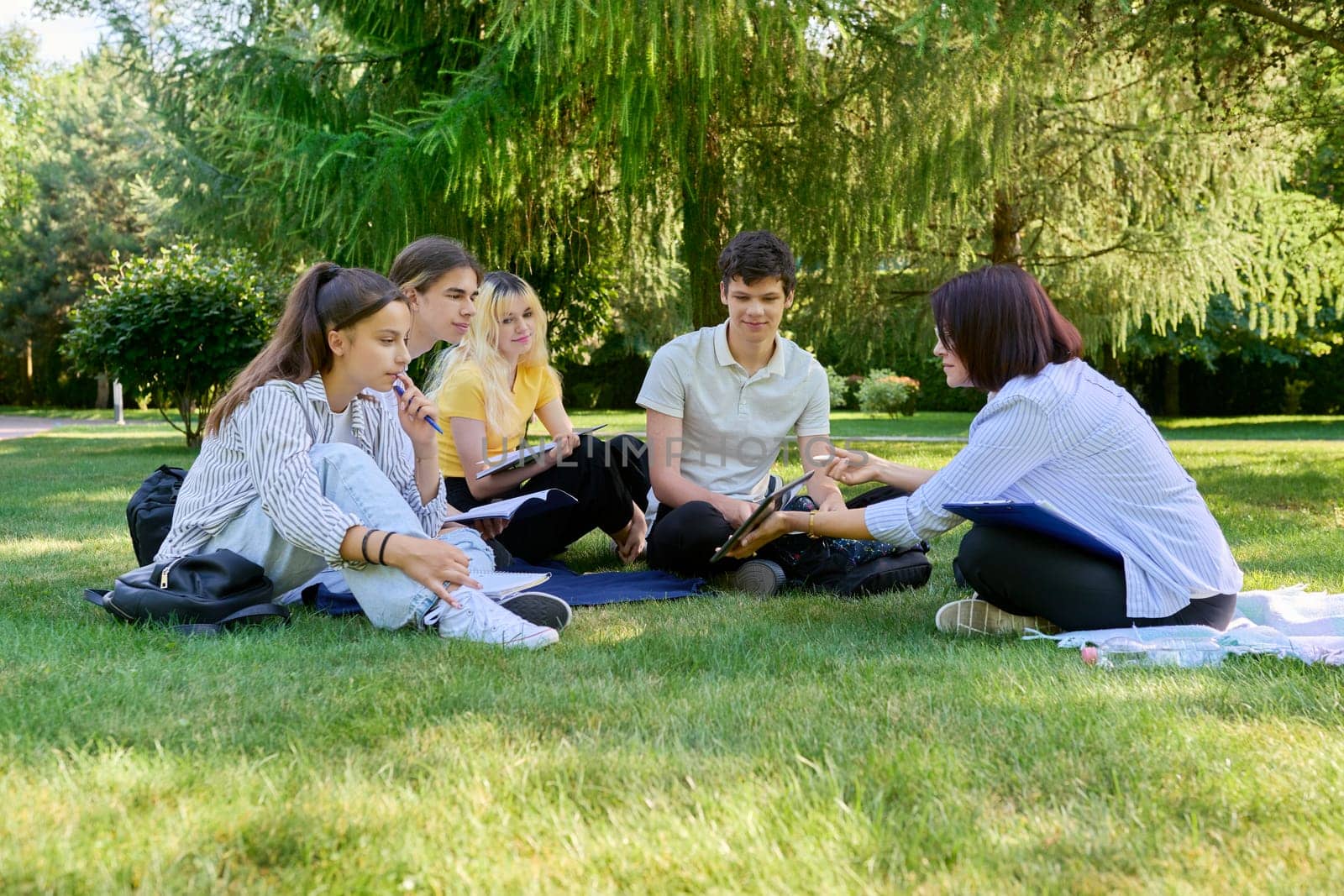 Outdoor, group of students with female teacher. Teenagers and mentor teacher talking sitting on grass in college park. Back to school, back to college, high school, education, teenagers concept