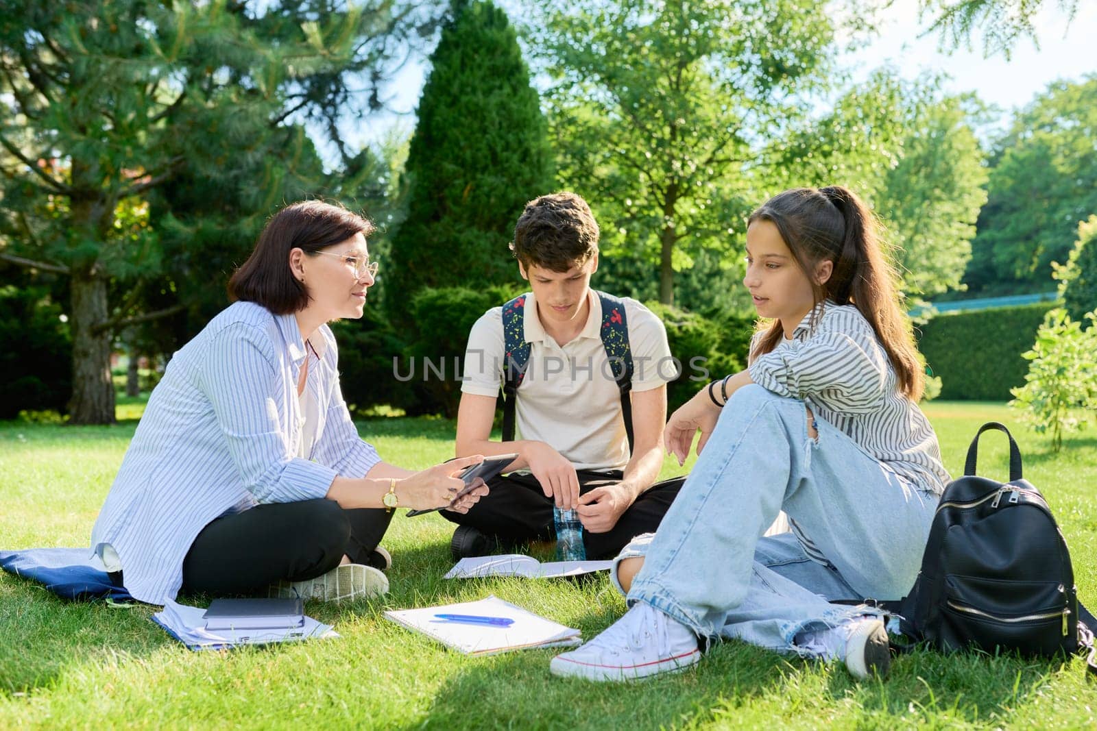 School teacher, psychologist, social worker talking to teenagers. Woman with teenage schoolchildren sitting on grass on campus lawn. Education, high school, psychology, mental health, adolescence