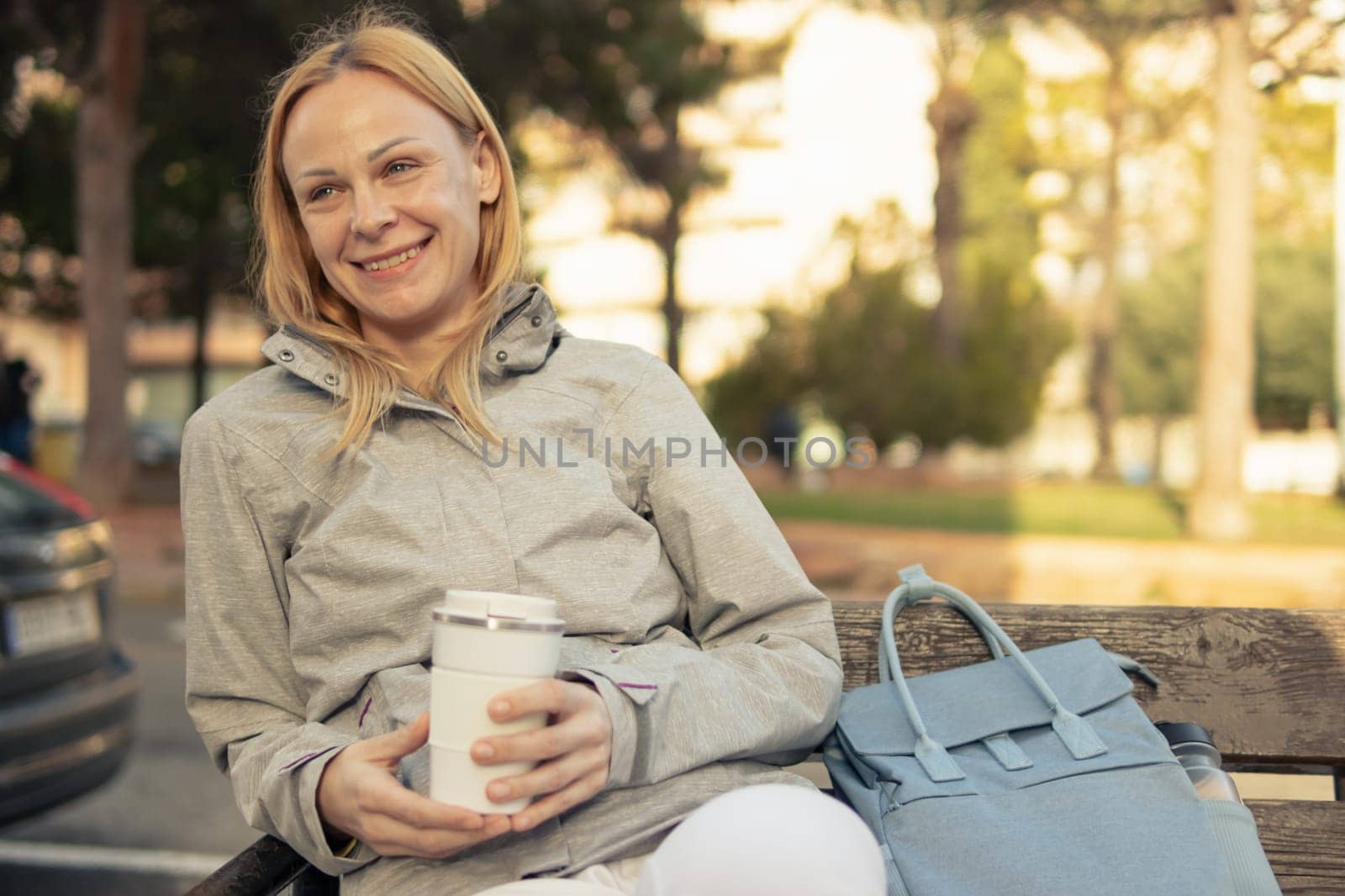 a smiling girl of European appearance with blond hair sits on a bench, drinks coffee from a white thermo mug, a girl sits in a gray jacket, a blue backpack lies nearby. High quality photo