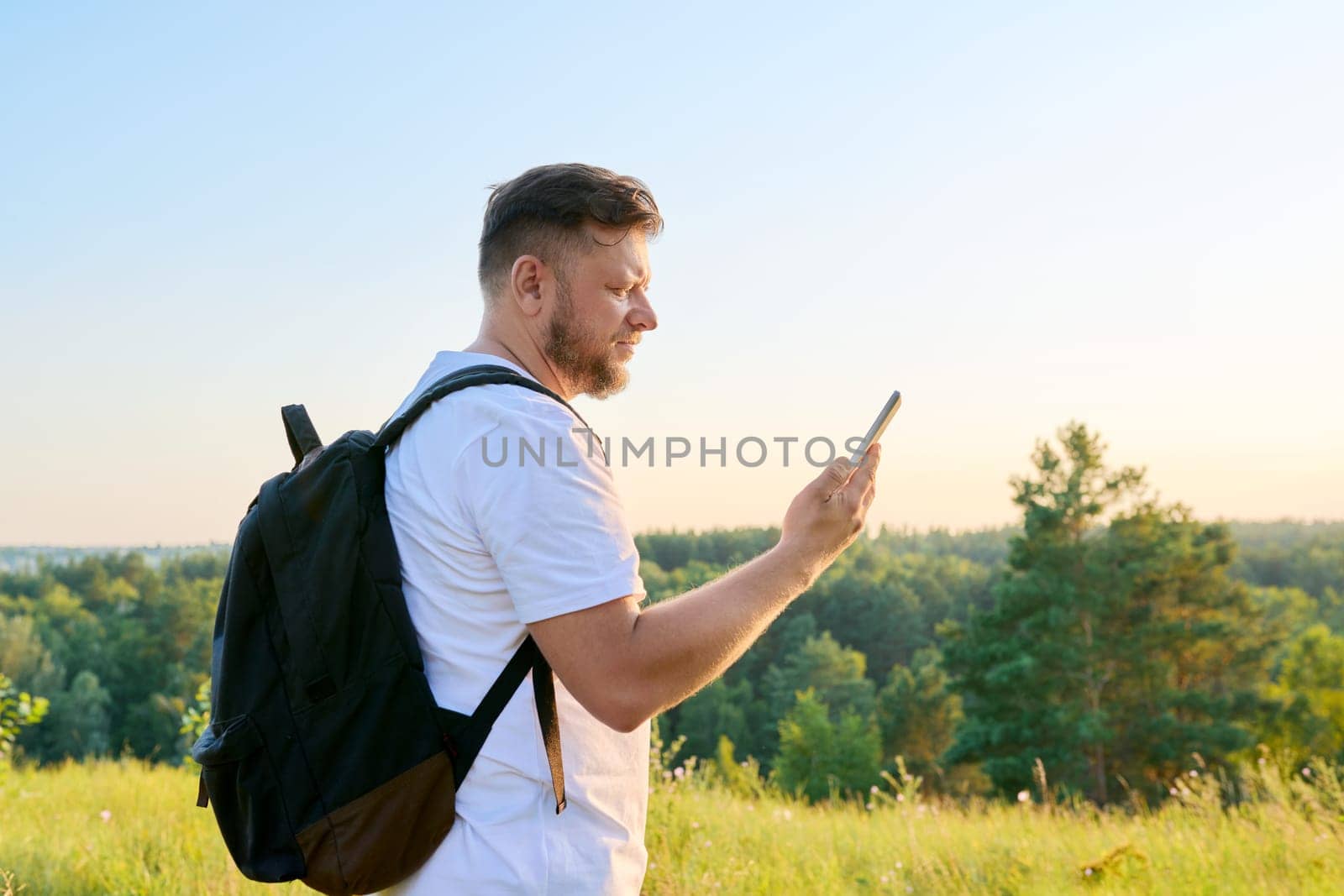 Middle aged bearded man with smartphone backpack on hike, making video call, talking looking at webcam. Wild summer nature meadow forest background. Leisure, technology, adventure, travel, people