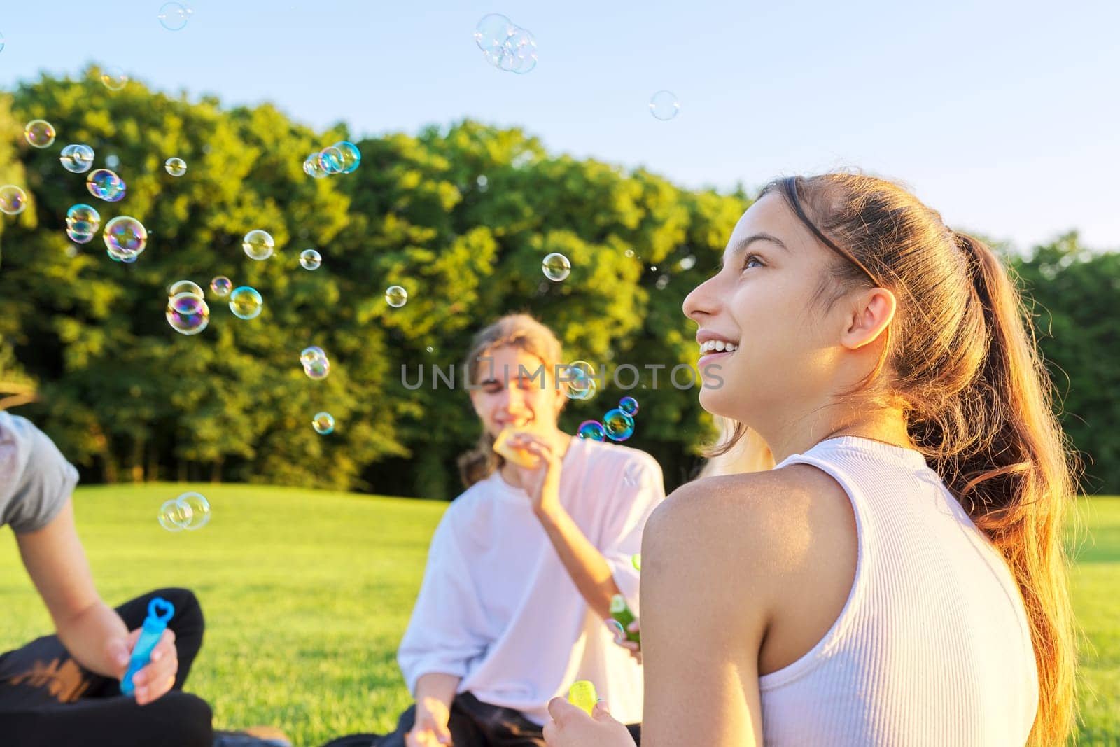 Happy teenage girl 13, 14 years old blowing bubbles. Teenager having fun with friends on lawn in park on sunny day. Fun, summer, vacation, friendship, adolescence concept