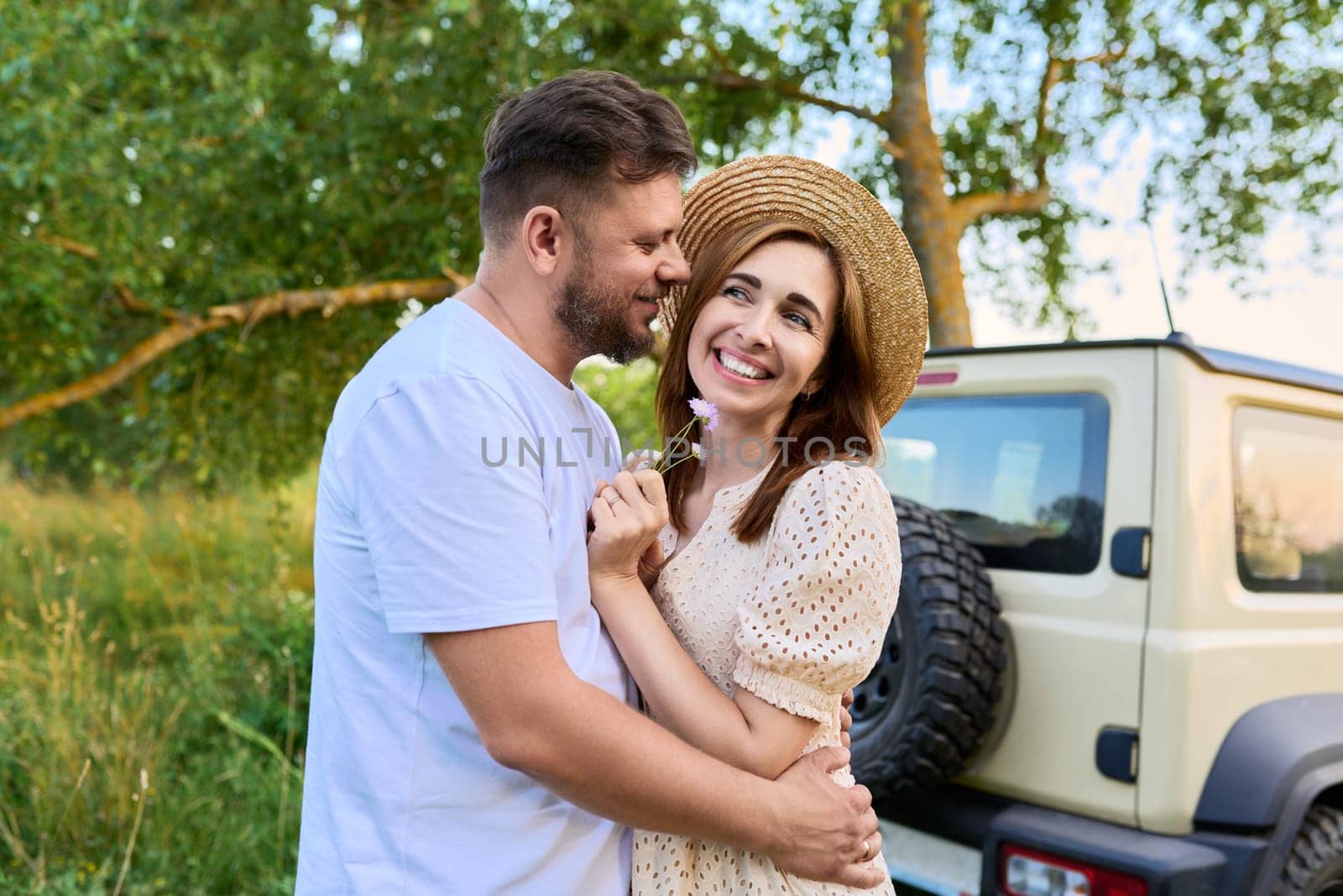 Love and romance of middle-aged couple. Happy hugging man and woman near car, summer nature wild meadow background. Relationship, holiday, date, trip, journey, weekend, family, people 40s concept