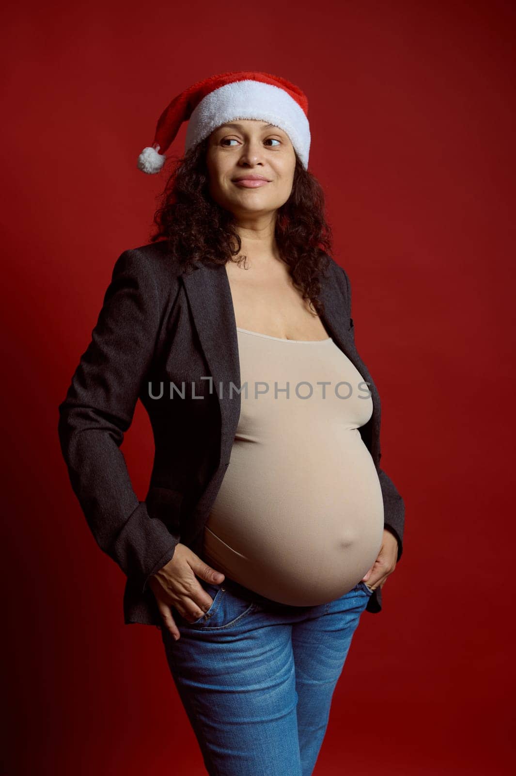 Studio portrait of emotional charismatic pregnant woman in Santa hat, smiling looking aside, isolated on red background by artgf