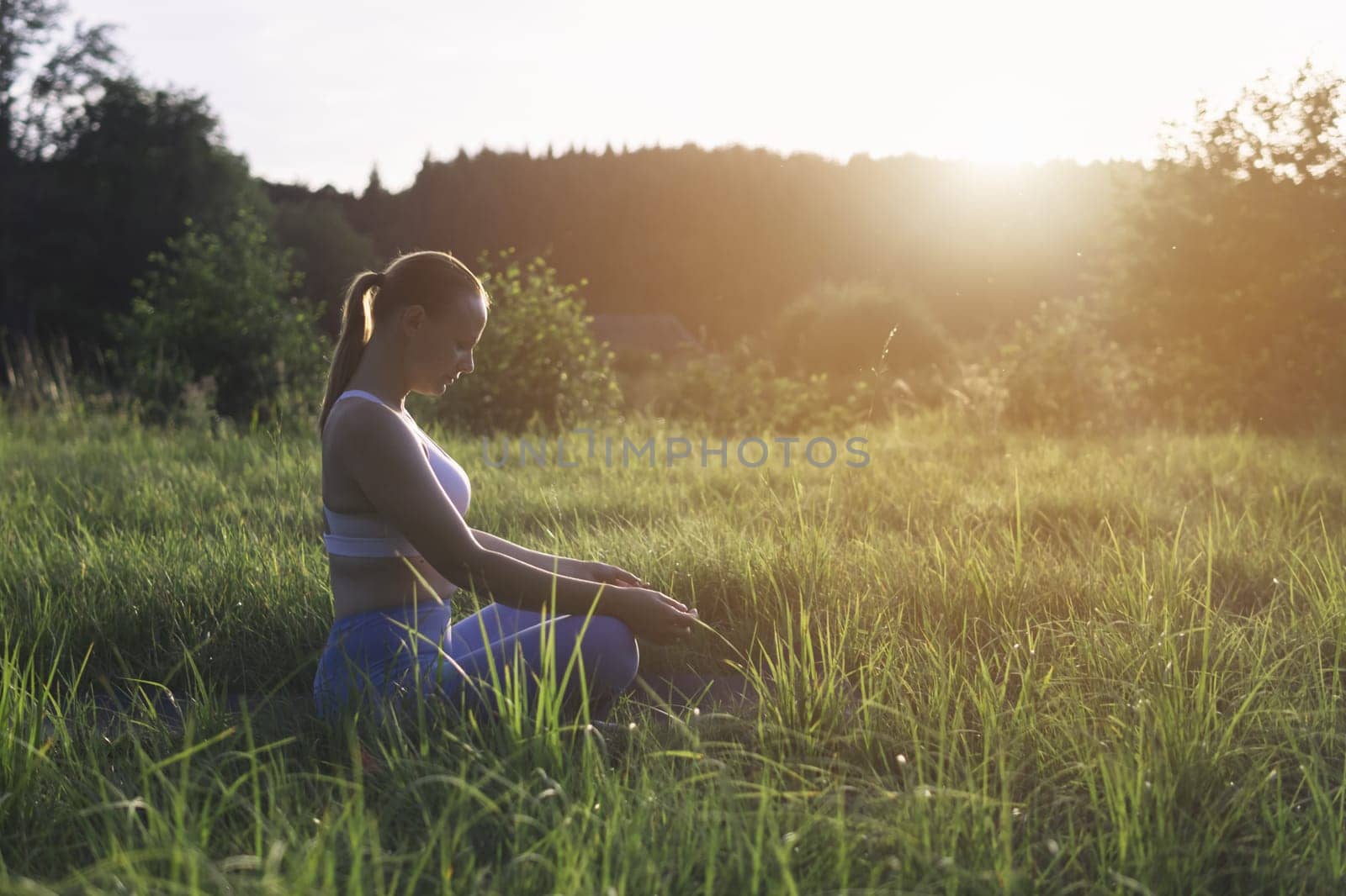 a girl of light appearance at sunset in a park doing exercises in sportswear, sitting on the grass, a silhouette of a woman, there is a place for an inscription. High quality photo
