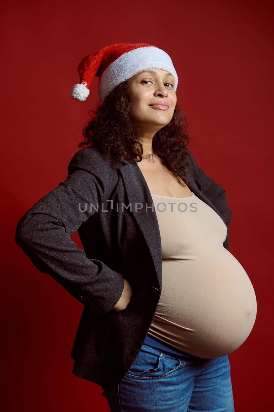 Studio portrait of emotional charismatic curly haired pregnant woman in stylish blazer, blue jeans and Santa hat, smiling cutely looking at camera, isolated red color backdrop. Happy New Year concept