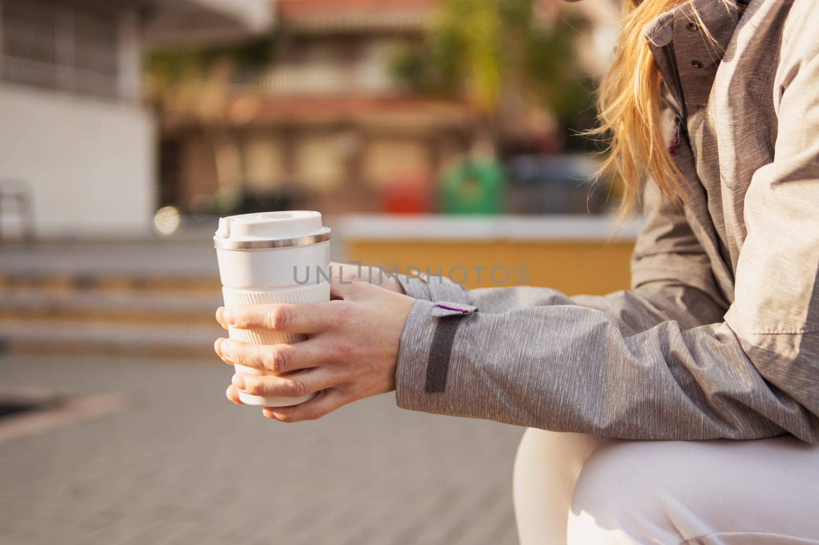 A girl is sitting on a bench in the park, holding a white thermo mug for tea or coffee in her hands. There is a place for an inscription, a close-up on her hands and a thermos. High quality photo
