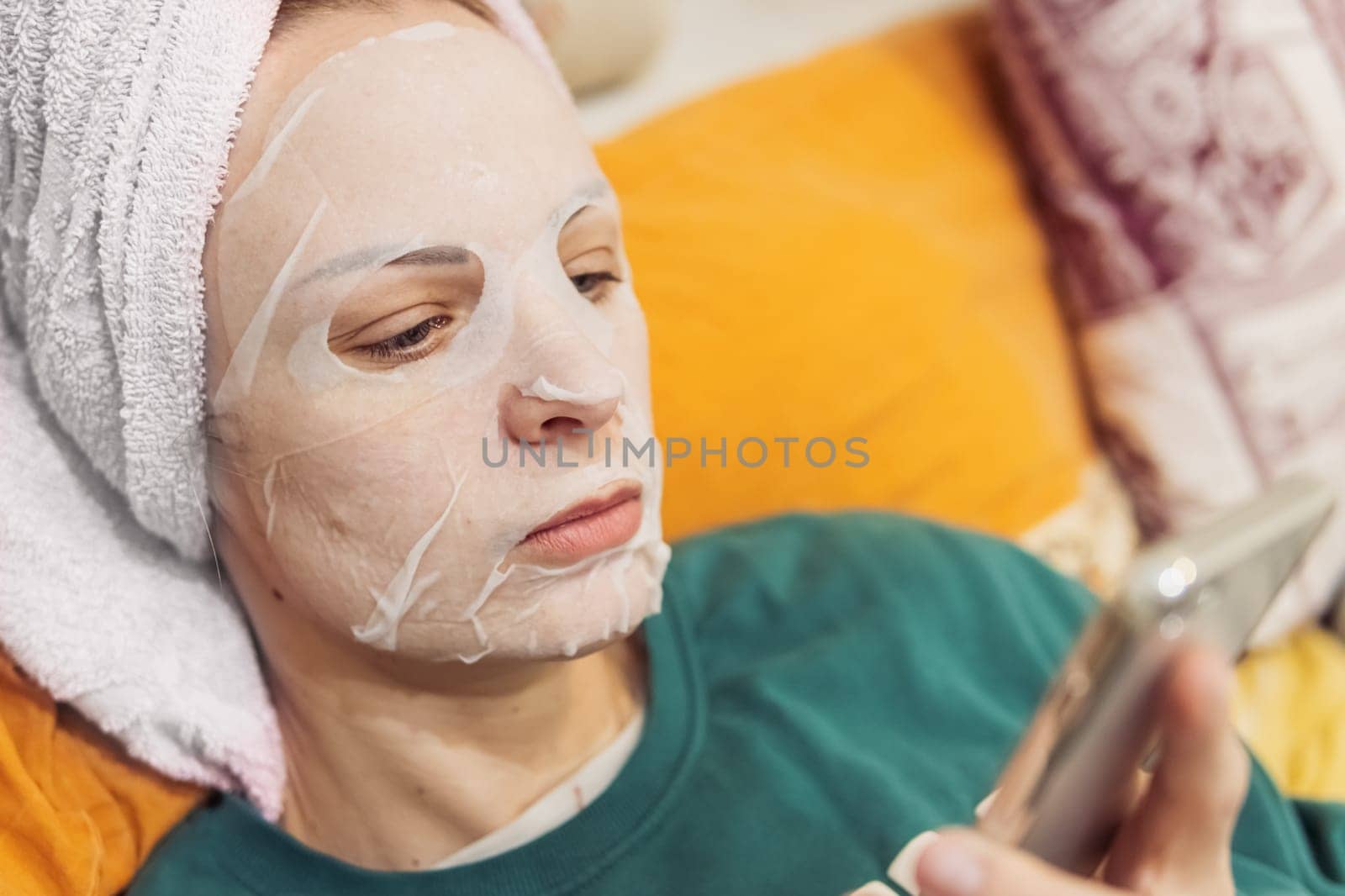 a girl of European appearance lies on a bed with a towel on her head and a mask on her face, doing procedures for facial rejuvenation, flipping through the phone. Relax for a girl. High quality photo