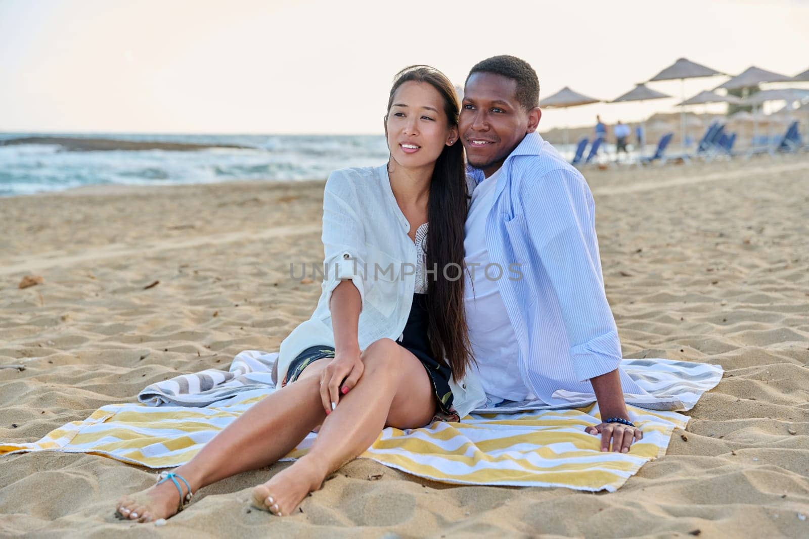 Happy multiethnic family resting on beach together. Asian woman and African man seated hugging each other on sand. Relationships, vacation travel and tourism at seaside resorts, people 20 30 years old