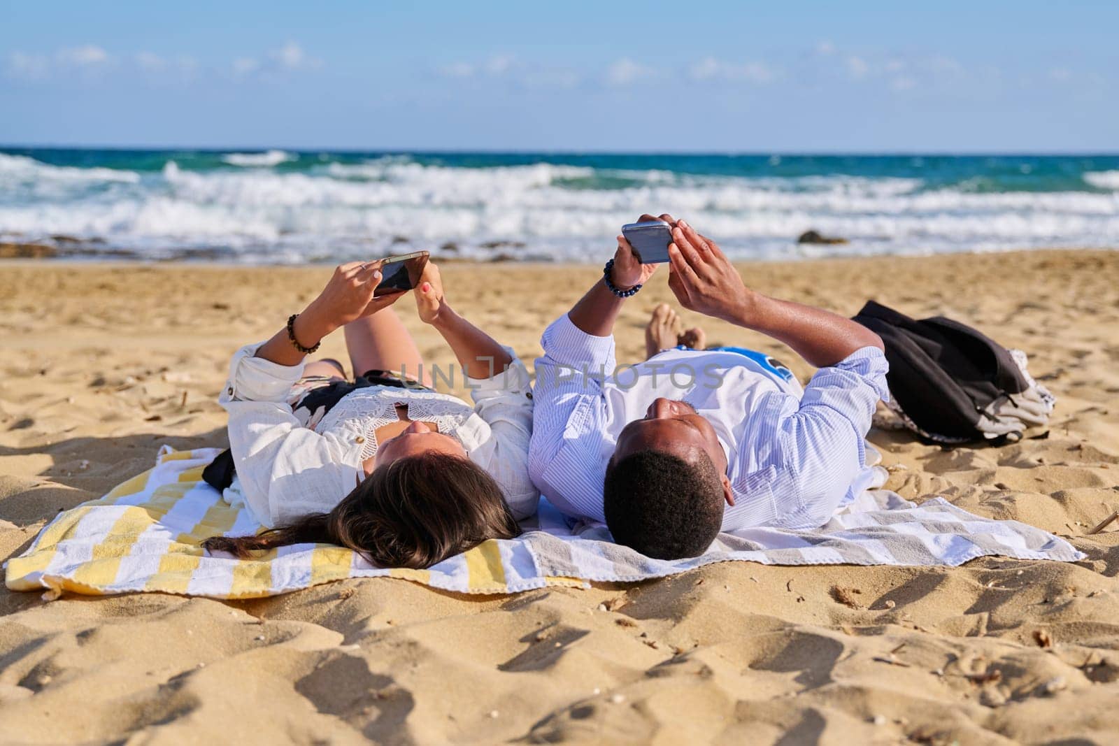 Young multicultural couple lying on beach using smartphones. Relaxing tourists lying on beach towels looking at smartphones. Lifestyle, technology, tourism, travel, nature, leisure, people concept