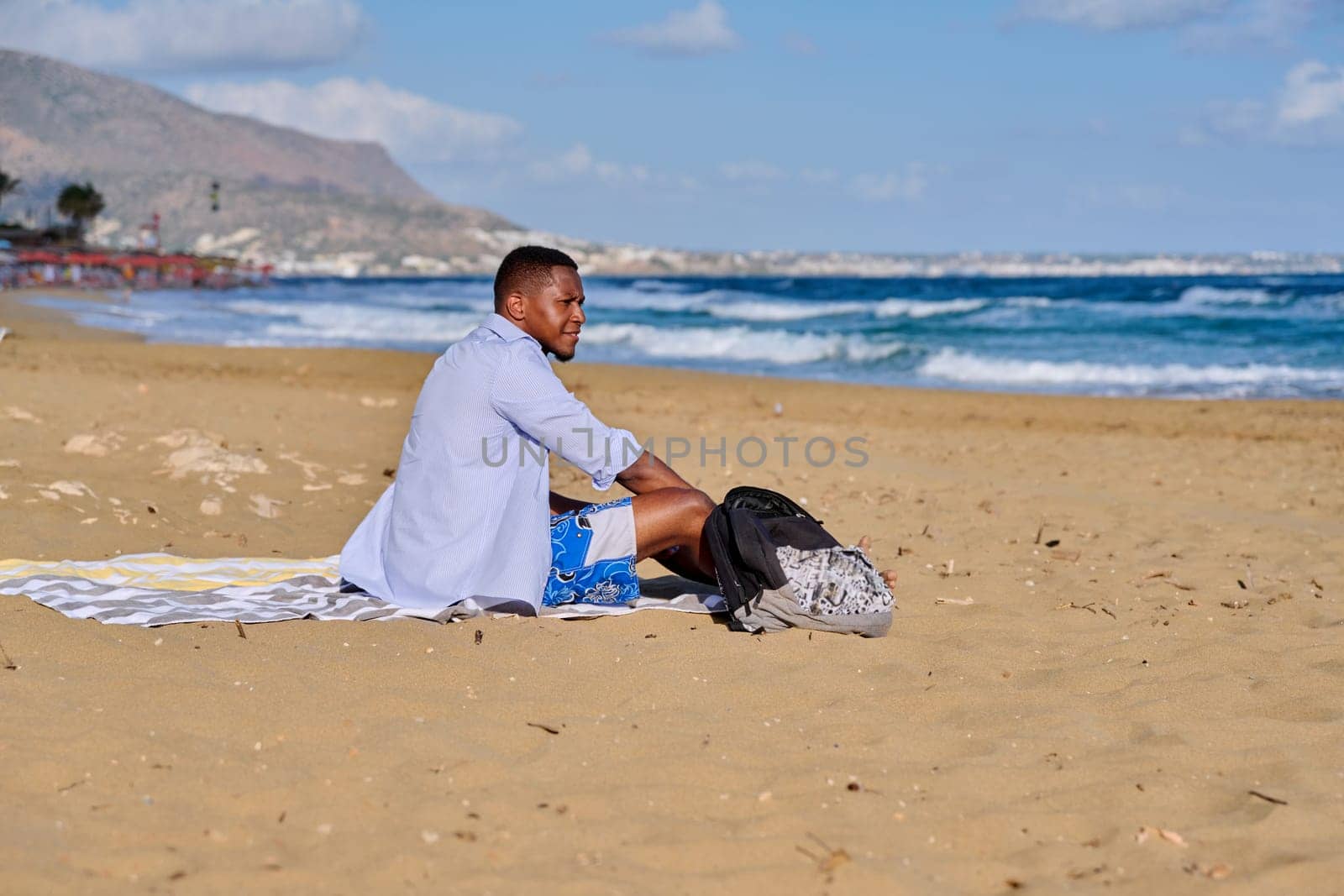 Young positive male tourist sitting resting on beach, copy space. Smiling African American male 30 years old sitting on sand, sea nature background. Lifestyle, travel, tourism, people concept