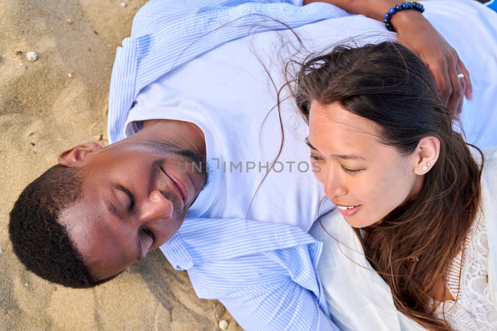 Young happy couple lying on the sand, top view. Loving multicultural couple, Asian woman and African man, faces close up. Love, relationship, beauty, romance, happiness, sensuality, people concept