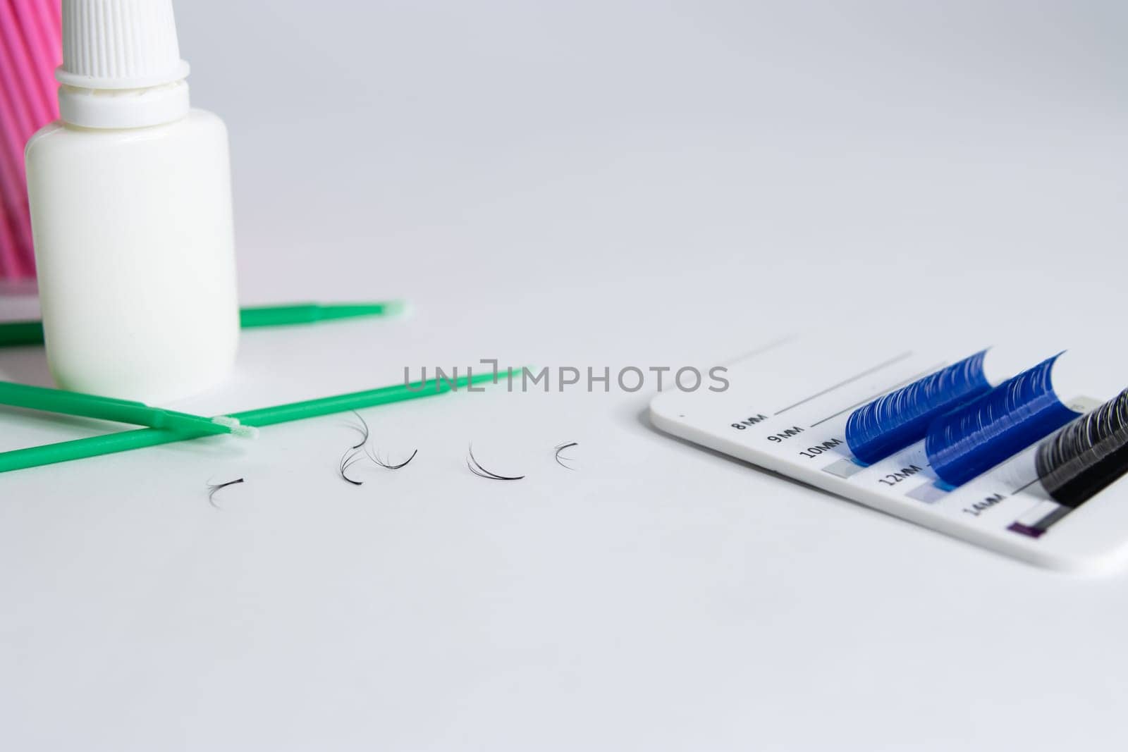 Artificial eyelashes in black and blue are pasted on a white tablet on a white background. On the left side are bundles of eyelashes, pink and green microbrushes and a white bottle of liquid.close-up