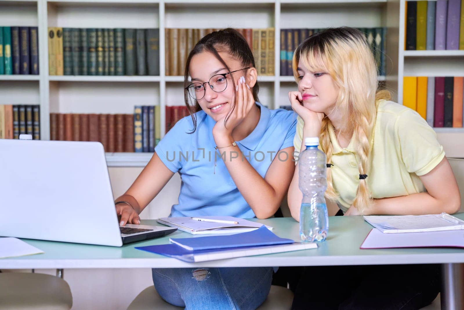 Two teenage schoolgirls students sitting with a laptop in the library, girls looking at laptop screen. Education, high school, technology, teamwork, learning, e-learning, adolescence concept