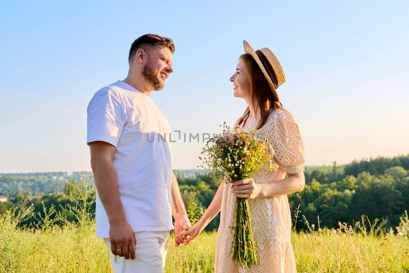 Couple holding hands smiling talking, summer nature blue sky background. Happy 40s age people, female with bouquet of flowers in dress hat, holiday, vacation, outdoor walk, relationship concept