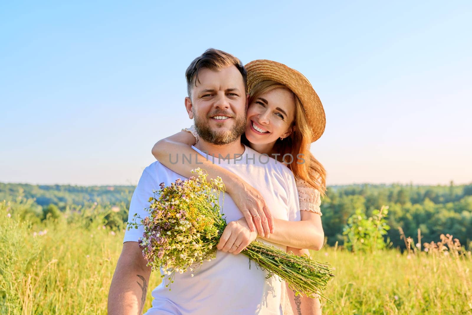 Happy middle-aged couple in love embracing, with bouquet of wildflowers, outdoors on summer nature meadow, looking at camera. Date, anniversary, family, wedding, celebration, relationship, 40s people