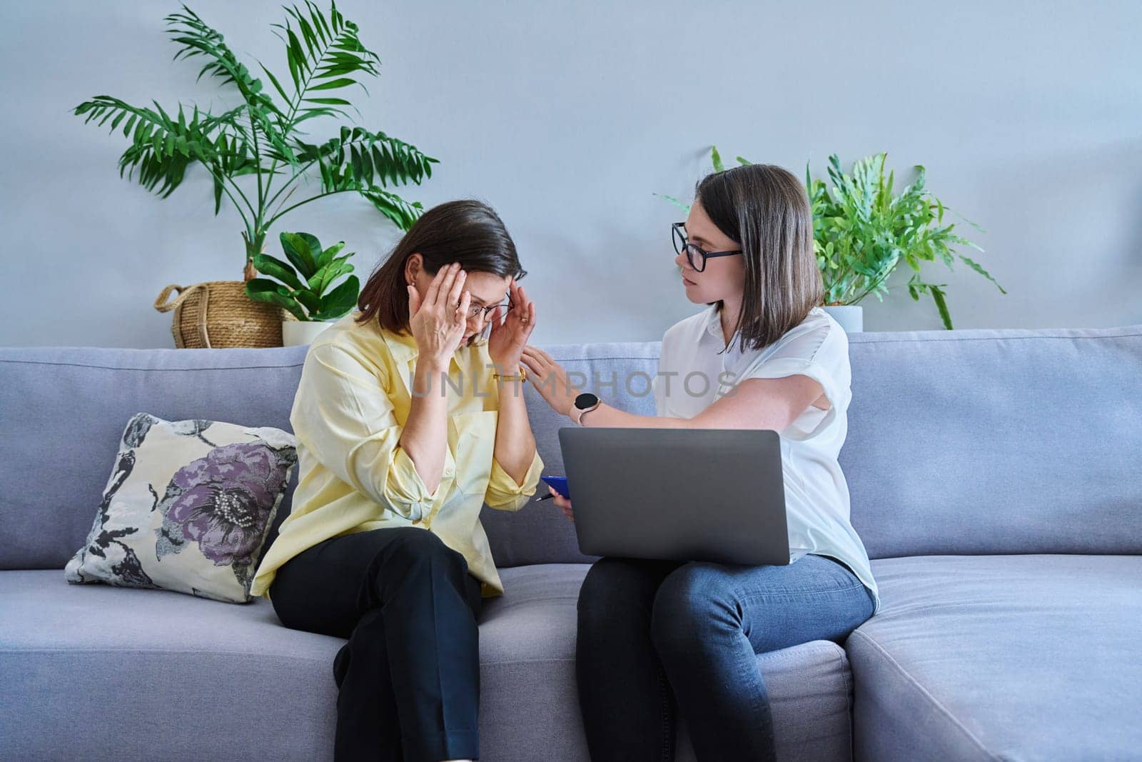 Female psychologist and middle aged woman patient sitting together on couch in office by VH-studio