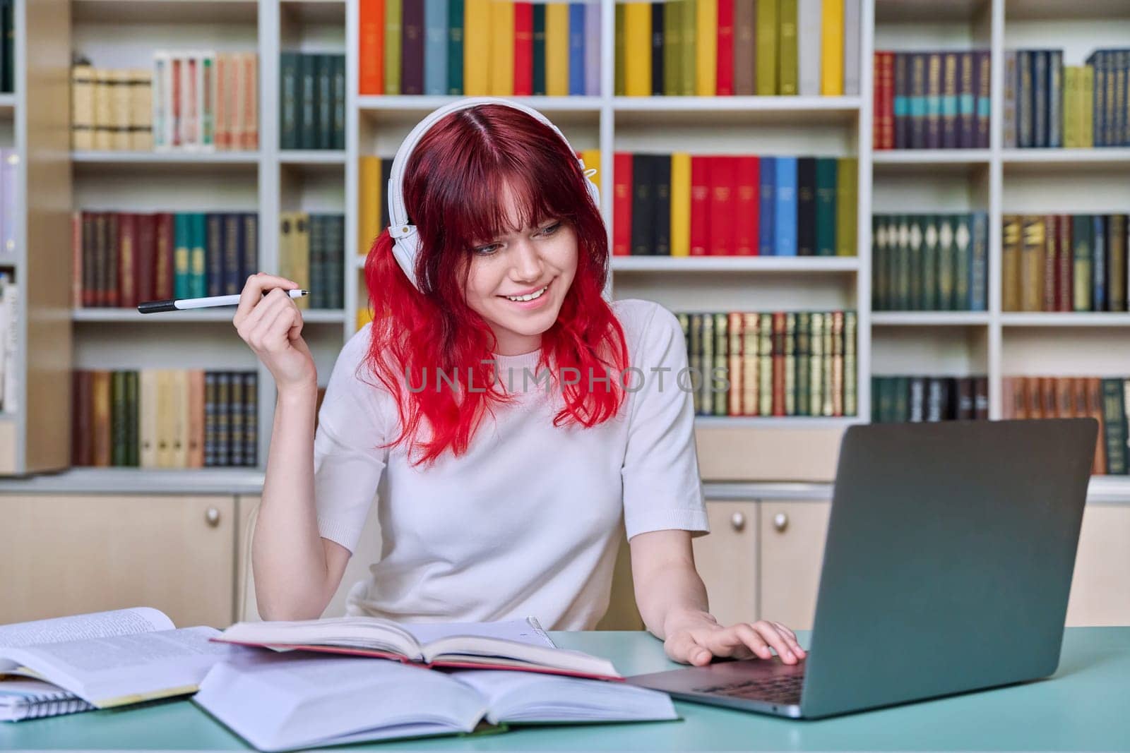 Teenage female student studying in library. Girl in headphones using laptop, looking at screen, watching webinar, making notes in notebook. Youth, technology, education, college, university concept